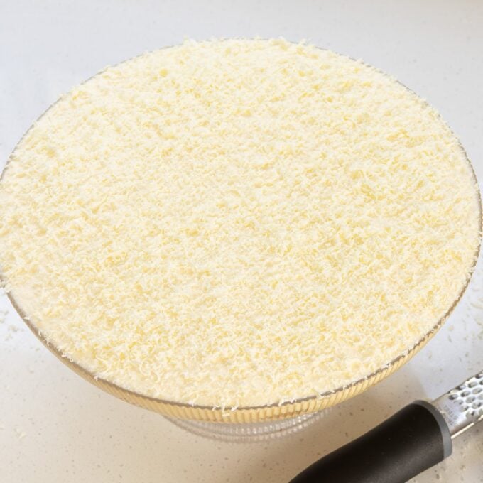 Limoncello Tiramisu A glass bowl filled with a dessert topped with a thick layer of finely grated white cheese, with a grater placed beside it.