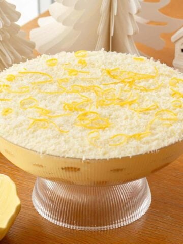 A glass bowl filled with a layered dessert topped with grated white chocolate and lemon zest, placed on a wooden table beside decorative white trees and a small house.