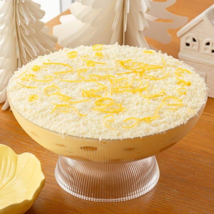 Limoncello Tiramisu A glass bowl filled with a layered dessert topped with grated white chocolate and lemon zest, placed on a wooden table beside decorative white trees and a small house.