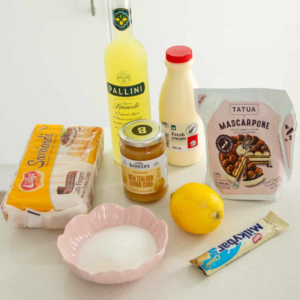 Limoncello Tiramisu Assorted dessert ingredients on a counter: ladyfingers, limoncello, fresh cream, mascarpone, lemon curd, a lemon, sugar, and a white chocolate bar.