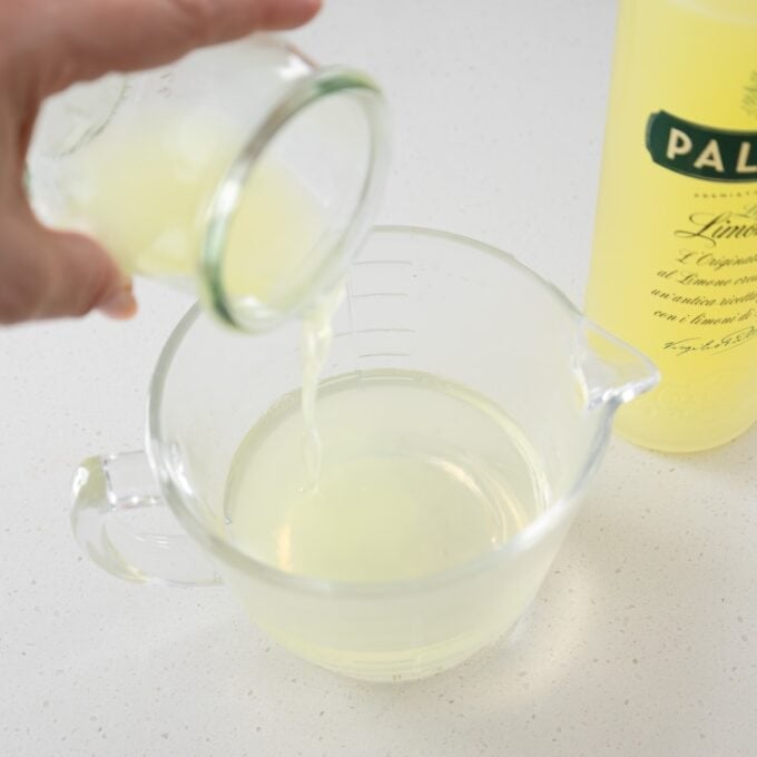 Limoncello Tiramisu Hand pouring a small glass of lime juice into a clear measuring cup, with a bottle of lime juice visible in the background on a white countertop.