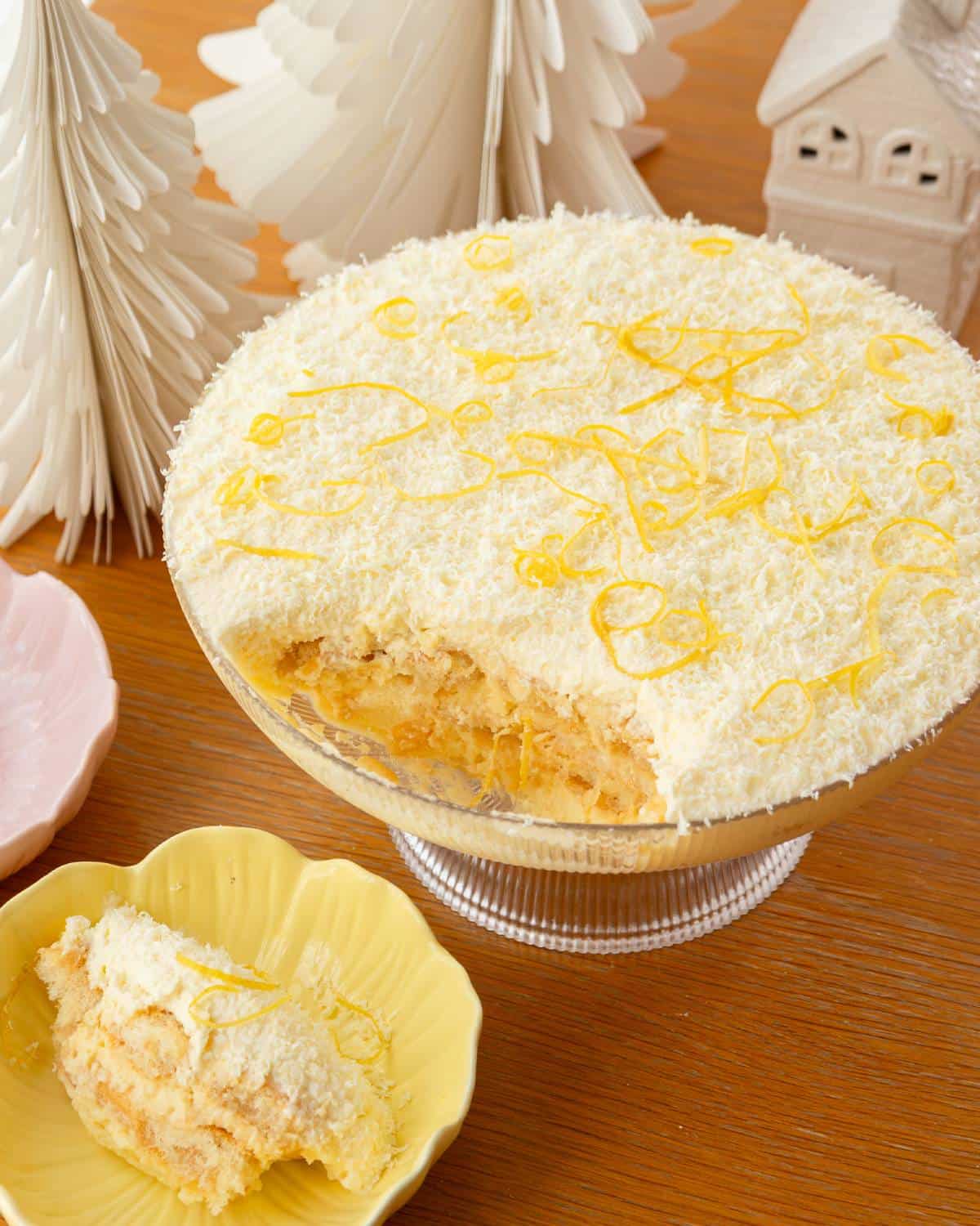 Limoncello Tiramisu A round dessert topped with white shredded coconut and honey is displayed with a portion served in a yellow bowl. Decorative paper trees and a house are in the background.