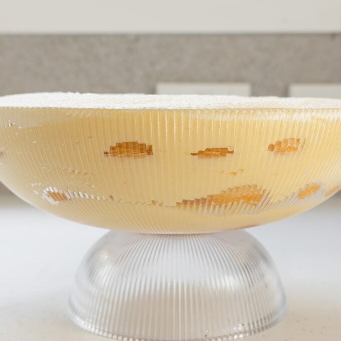 Limoncello Tiramisu A glass bowl filled with layered yellow custard dessert, visible ladyfinger pieces, and a dusting of powdered sugar on top, resting on a white surface.