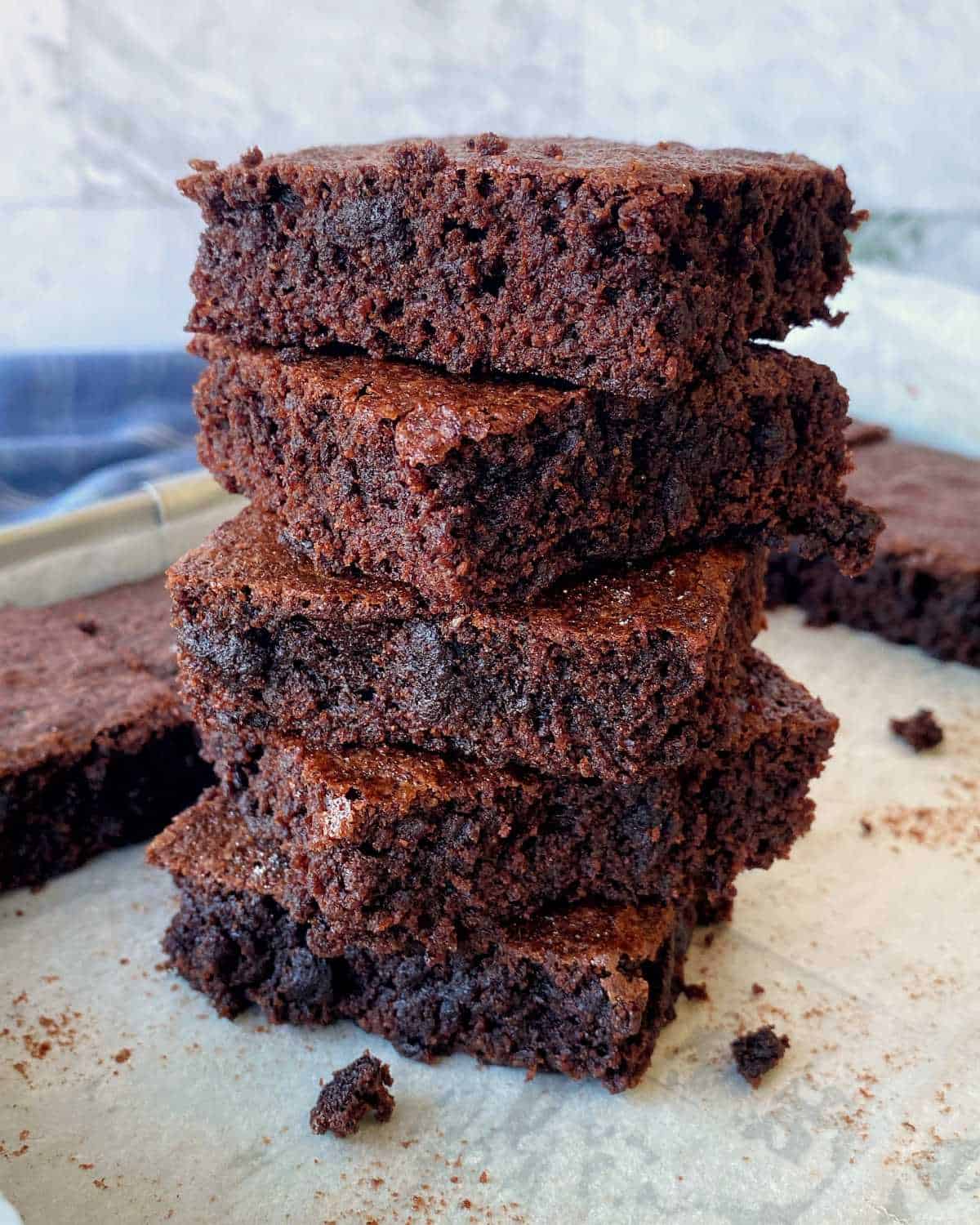 A stack of four thick, fudgy brownies sits on parchment paper, with more brownies and crumbs scattered in the background.