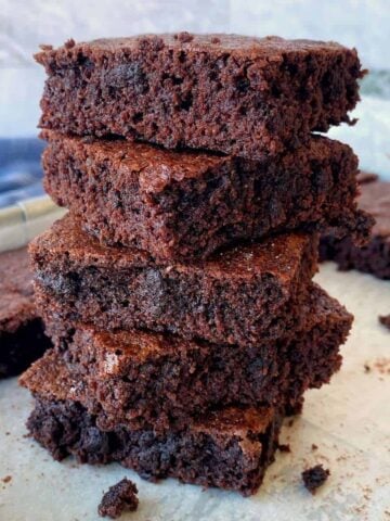 A stack of four chocolate brownies on parchment paper, with more brownies in the background.