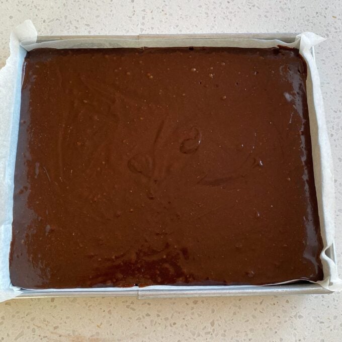 Big One Bowl Chocolate Brownie A rectangular baking tray lined with parchment paper, filled with raw chocolate brownie batter, sits on a light-colored countertop.