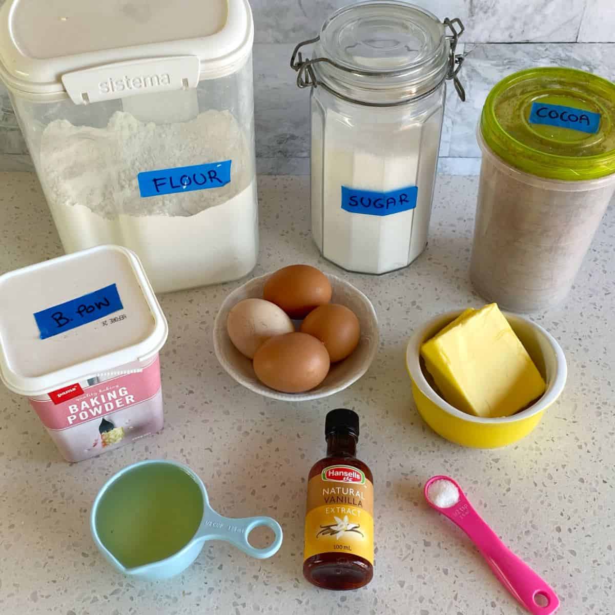 Big One Bowl Chocolate Brownie A variety of baking ingredients on a countertop, including flour, sugar, cocoa, baking powder, eggs, butter, vanilla extract, a measuring cup of liquid, and a pink spoon of salt.