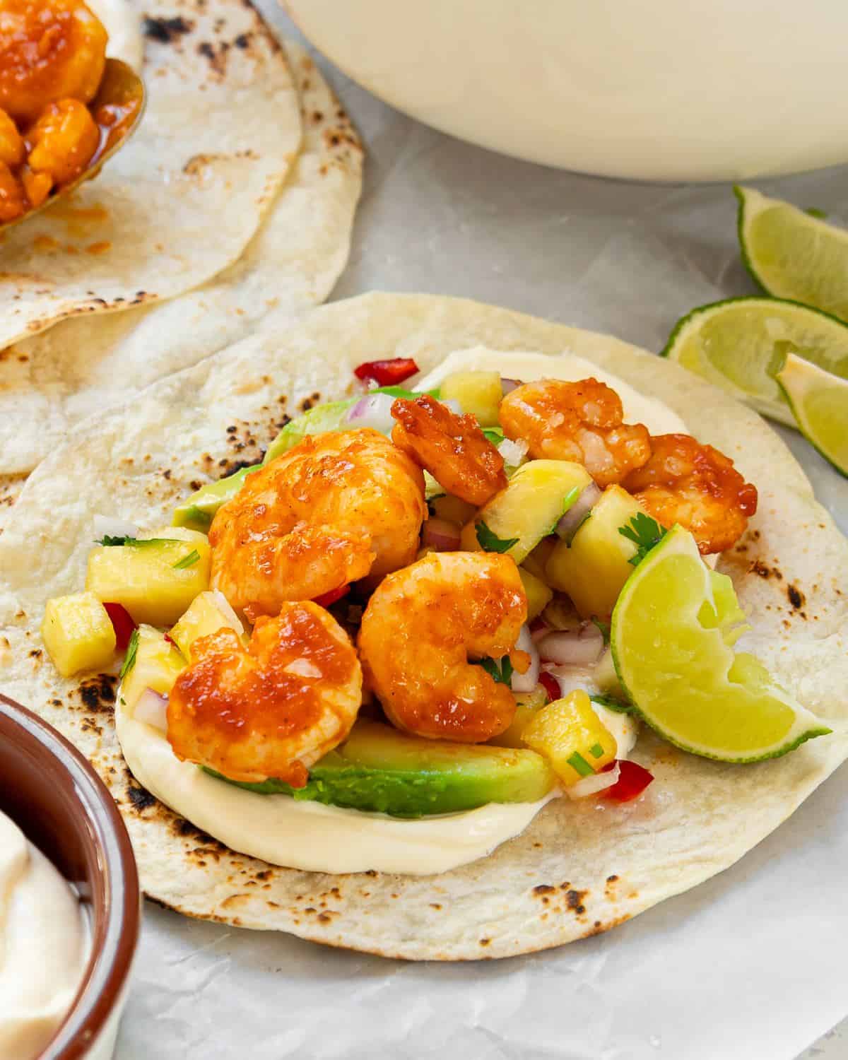 A corn tortilla topped with cooked shrimp, diced pineapple, avocado slices, chopped herbs, and a lime wedge, with sauce spread underneath and lime wedges on the side.