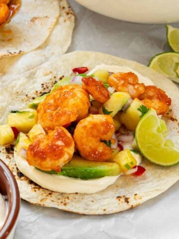 A soft tortilla topped with shrimp, avocado, pineapple salsa, mayonnaise, and lime wedges on the side.