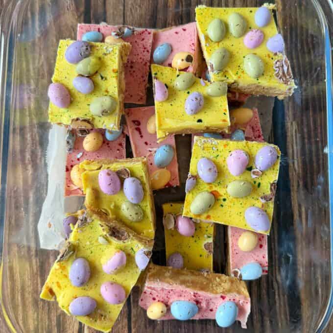 Speckled Mini Egg Slice A glass dish containing colorful dessert bars topped with yellow and pink icing and decorated with pastel candy eggs, set on a wooden surface.