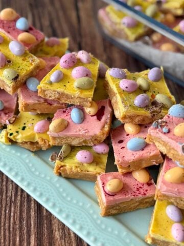 Colorful dessert bars topped with pink and yellow frosting and chocolate candy eggs are stacked on a blue plate, with more bars visible in a glass dish behind them.