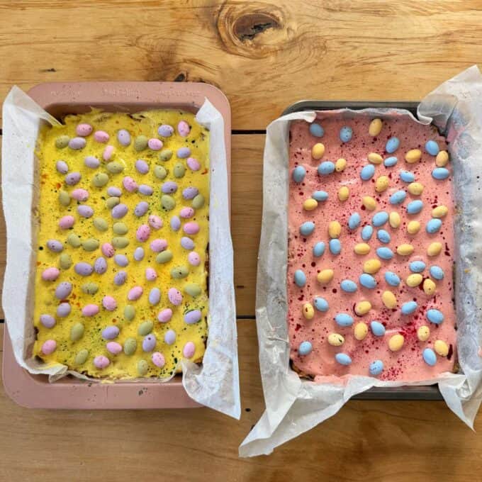 Speckled Mini Egg Slice Two baking trays lined with parchment paper hold yellow and pink tray bakes topped with pastel-colored candy eggs on a wooden surface.