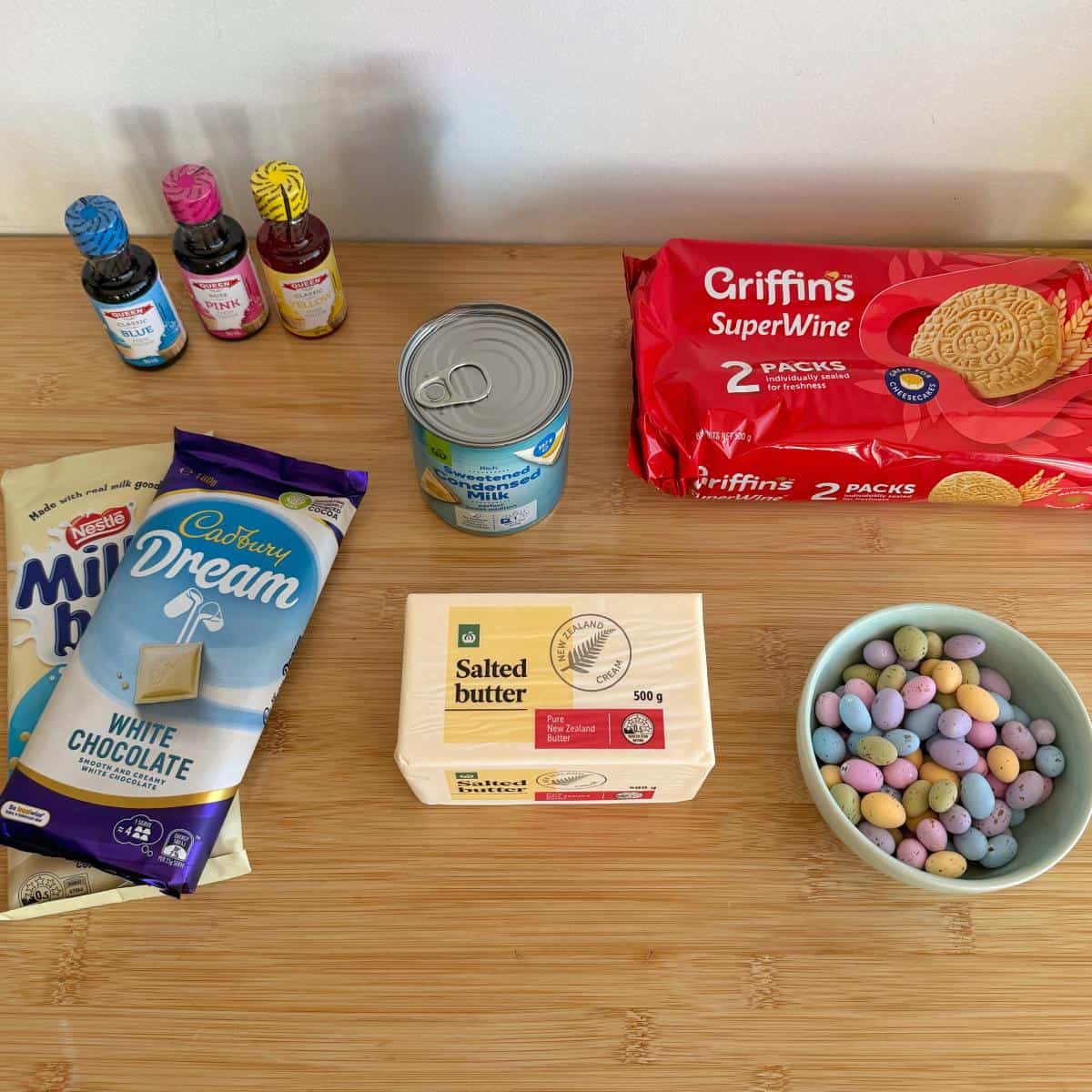 Speckled Mini Egg Slice Baking ingredients on a wooden surface, including food coloring, canned condensed milk, white chocolate, salted butter, Super Wine biscuits, and a bowl of candy-coated chocolates.