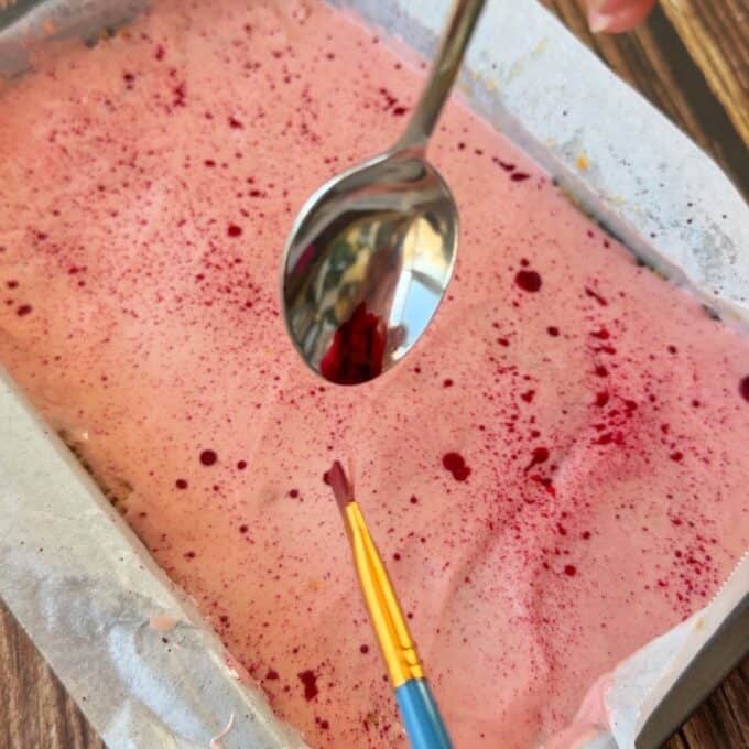 Speckled Mini Egg Slice A tray of pink frosted dessert is being decorated with red splatters using a paintbrush, while a spoon is held above the surface.