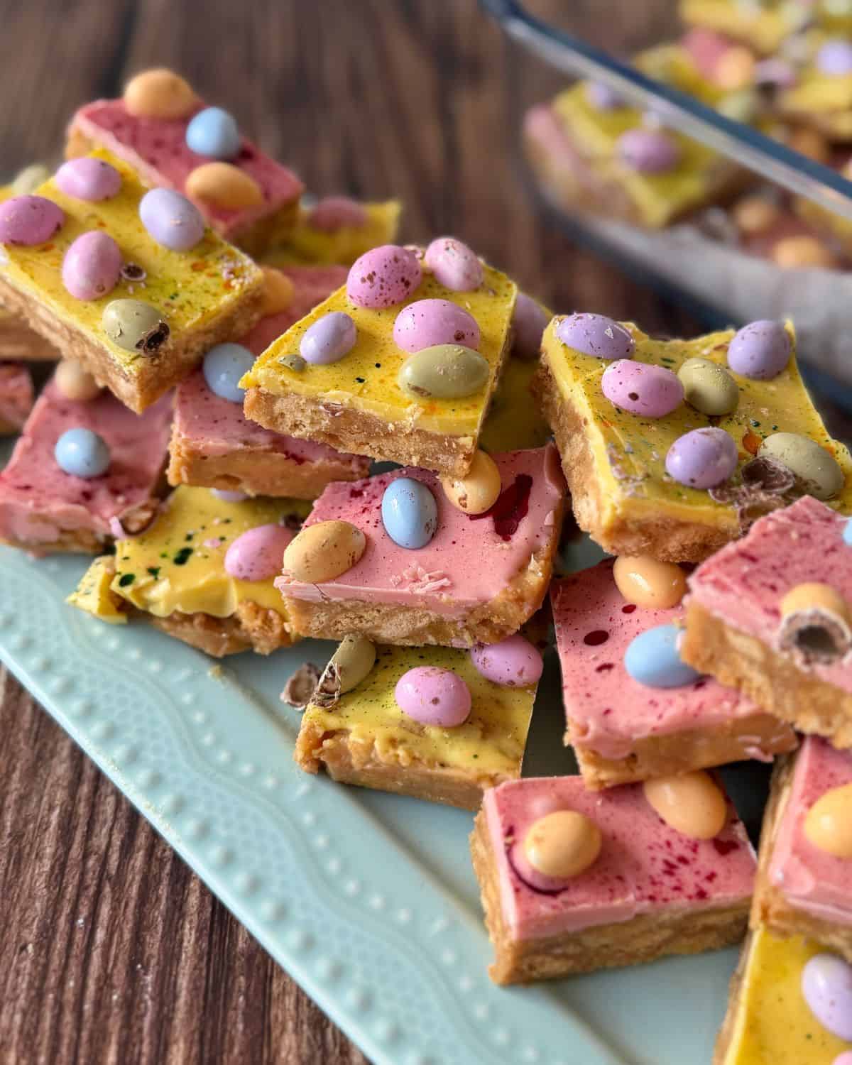 Speckled Mini Egg Slice Colorful dessert bars topped with pastel candy eggs are stacked on a light blue plate, with more bars visible in a glass dish in the background.