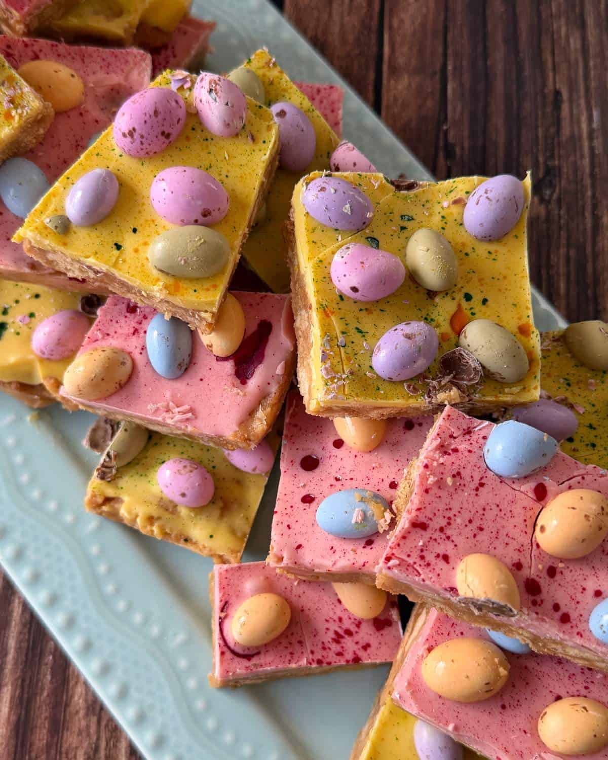 Rectangular pieces of pink and yellow chocolate bark topped with pastel candy-coated chocolate eggs are arranged on a white plate.