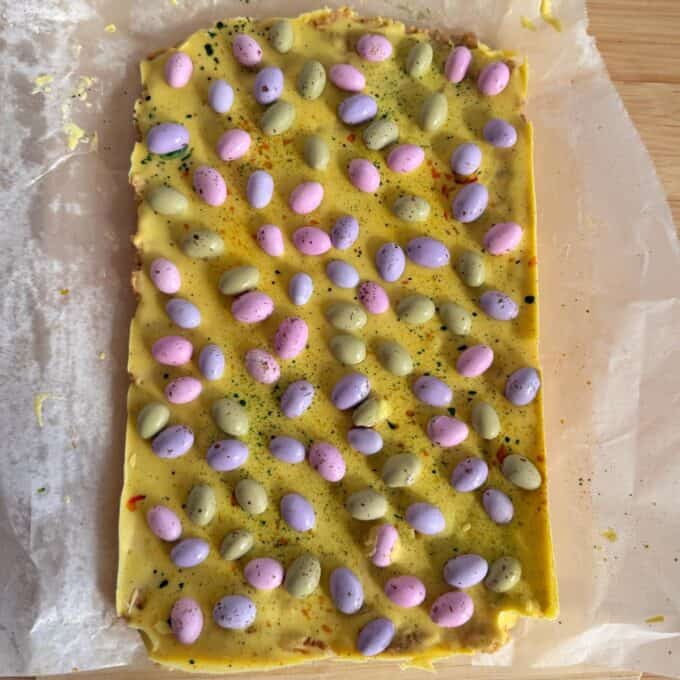 Speckled Mini Egg Slice A slab of yellow chocolate bark topped with pastel-colored candy-coated chocolate eggs, set on parchment paper on a wooden surface.