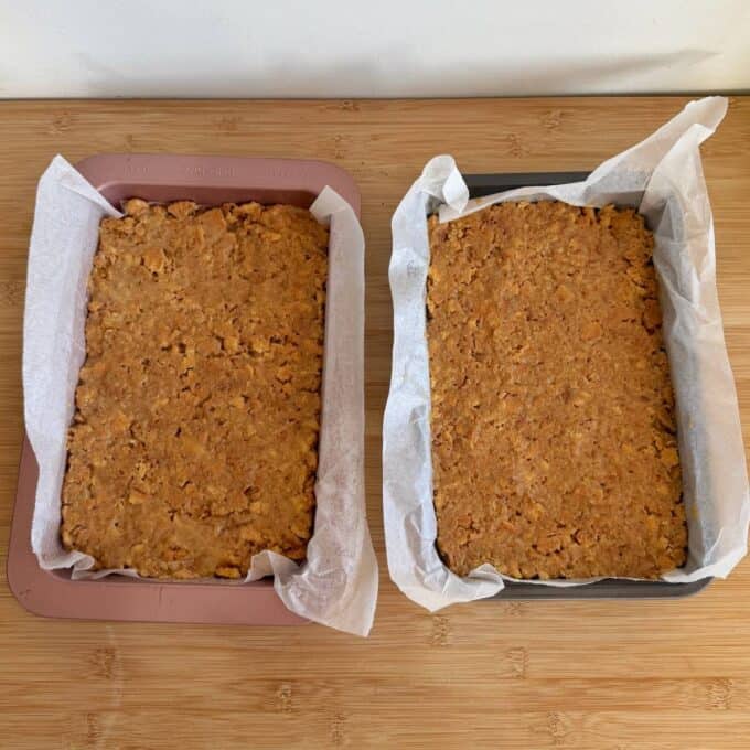 Speckled Mini Egg Slice Two rectangular baking trays lined with parchment paper, each filled with an unbaked, crumbly orange-brown mixture, are placed on a wooden surface.