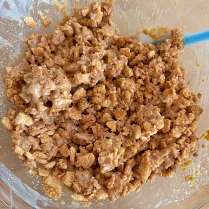 Speckled Mini Egg Slice A close-up of a mixing bowl filled with a mixture of crushed cereal and a sticky, light brown coating, with a blue spatula partially visible on the right.