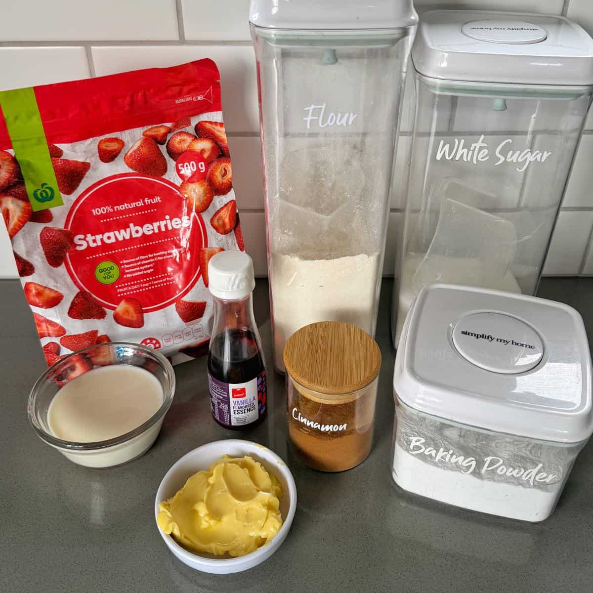 Strawberry Cobbler Baking ingredients on a countertop including flour, white sugar, baking powder, cinnamon, milk, butter, vanilla extract, and a bag of dried strawberries.