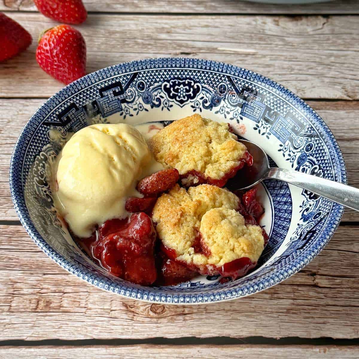 Strawberry Cobbler A bowl with strawberry cobbler and a scoop of vanilla ice cream, served with a spoon on a blue patterned plate; fresh strawberries are in the background.
