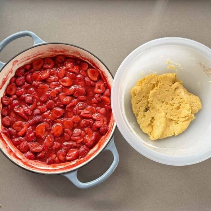 Strawberry Cobbler A pot of cooked strawberries in syrup sits next to a bowl of uncooked dough on a gray countertop.