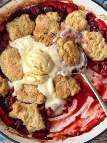 A baked strawberry cobbler in a round dish, topped with a scoop of melting vanilla ice cream and a spoon resting inside.