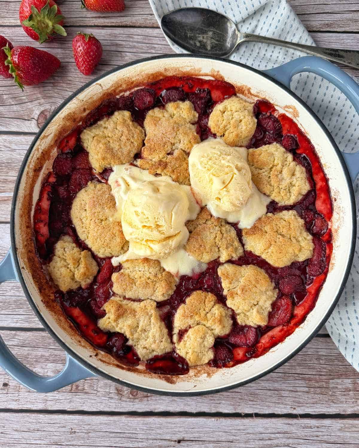 Strawberry Cobbler A baked strawberry cobbler topped with two scoops of melting vanilla ice cream in a round dish, with fresh strawberries and a spoon nearby.
