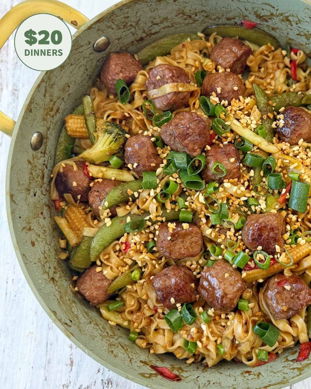 A skillet filled with noodles, meatballs, mixed vegetables, chopped green onions, and crushed peanuts, with a “$20 dinners” label in the corner.