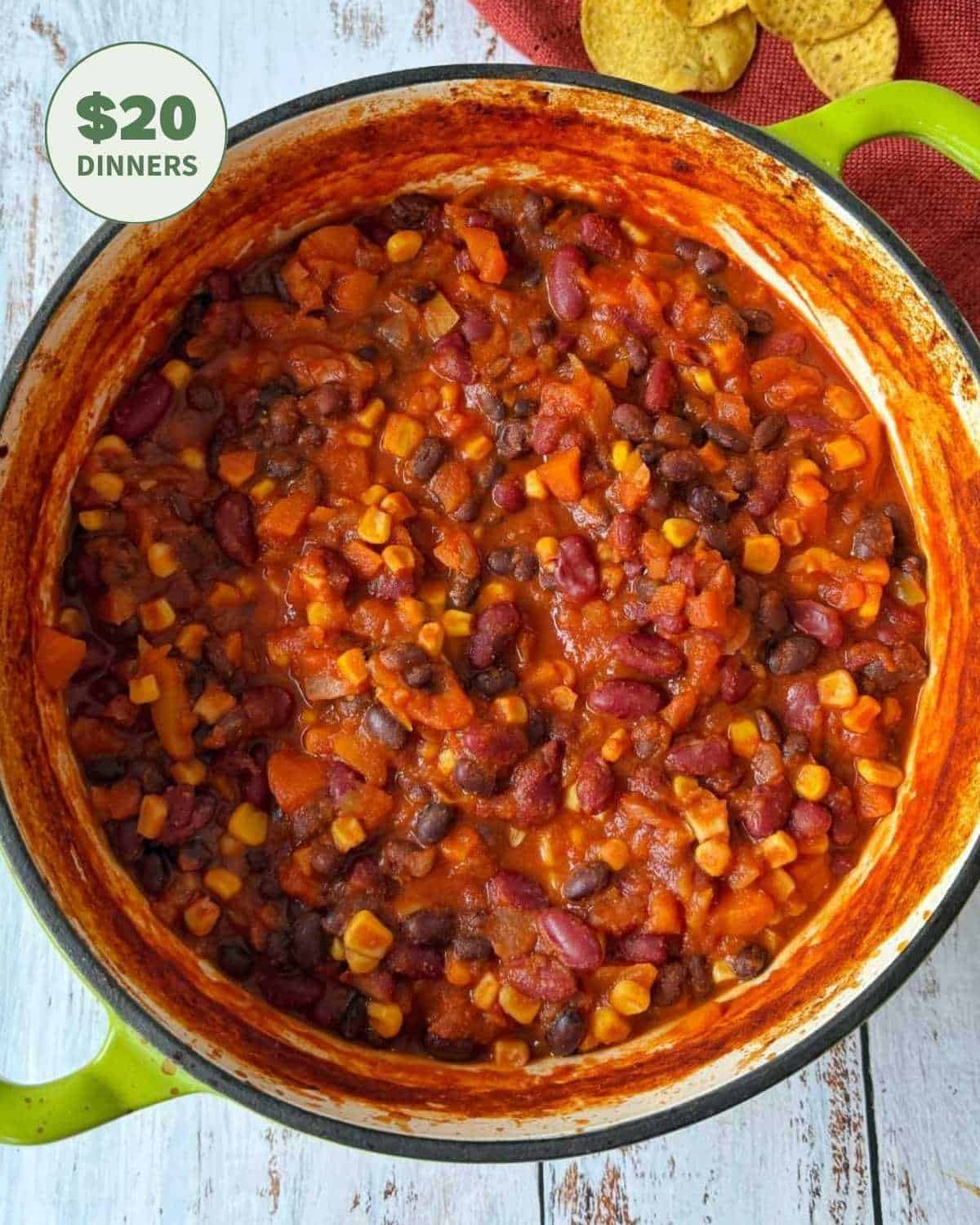 A large pot filled with vegetarian chili made of beans, corn, carrots, and tomato sauce, with some tortilla chips visible in the background. A $20 DINNERS label appears in the corner.