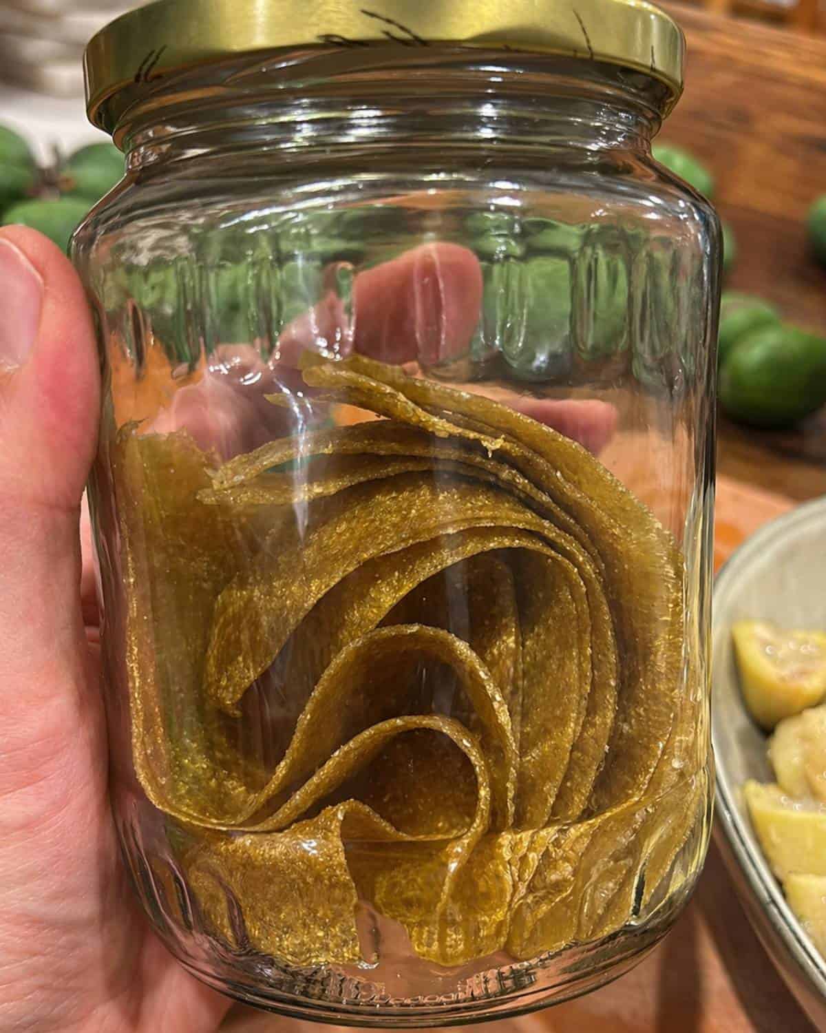 Feijoa Leather A hand holds a clear glass jar filled with rolled sheets of yellowish fruit leather, with a gold lid on top. Green fruits and a plate are visible in the blurred background.