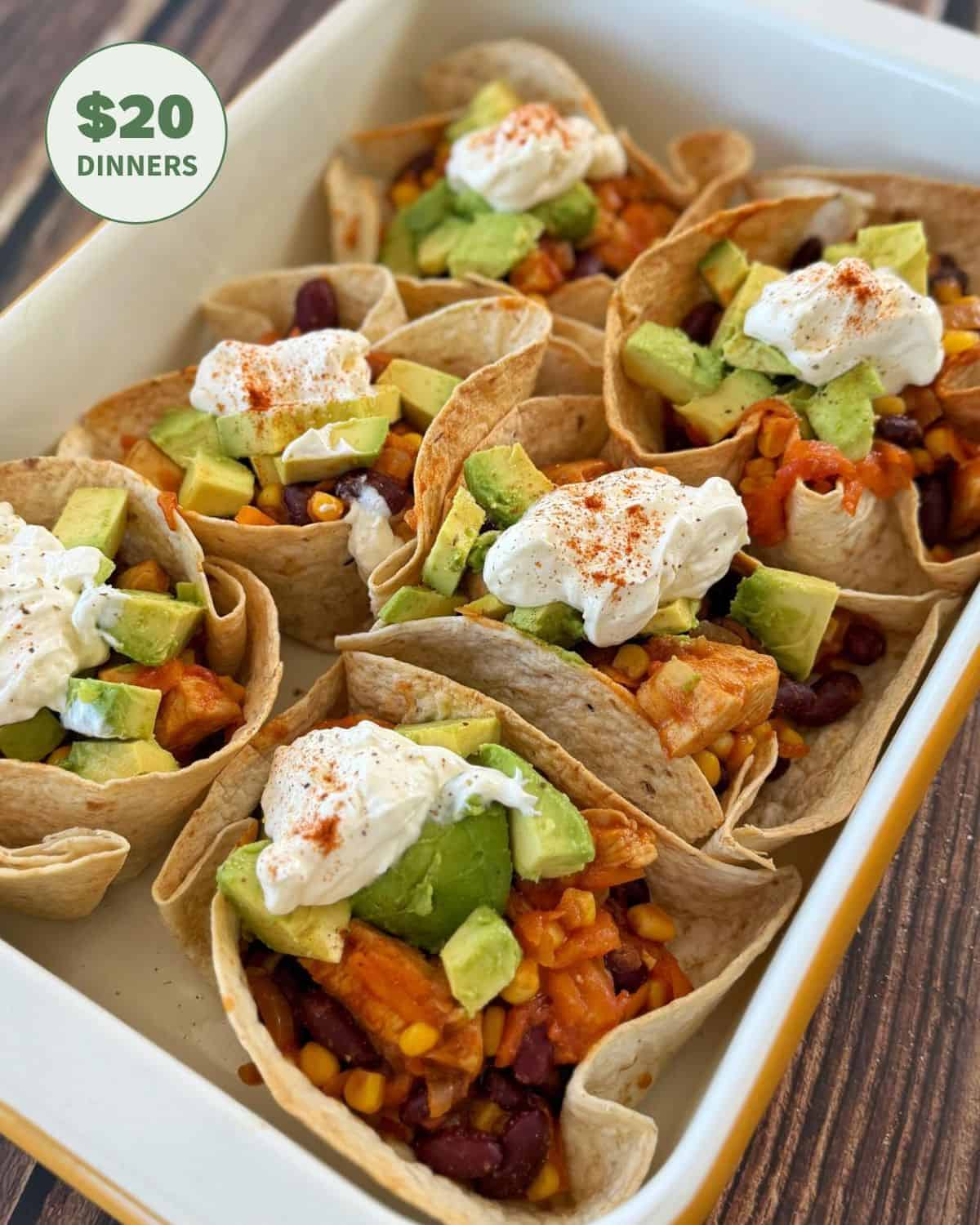 A baking dish with six tortilla cups filled with beans, corn, rice, avocado, and topped with sour cream and paprika. A graphic reads $20 Dinners in the corner.