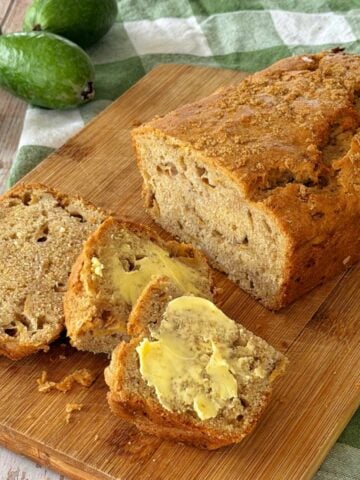 A Feijoa Loaf with three slices cut, one spread with butter, sits on a wooden board. Feijoa fruits and a green checkered cloth add a fresh touch in the background.