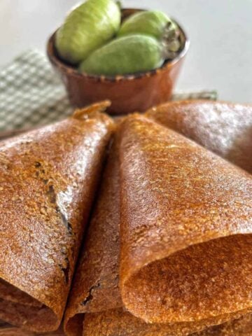 Several rolled brown dosa or crepes are arranged on a plate, with a bowl of green guavas placed in the background.