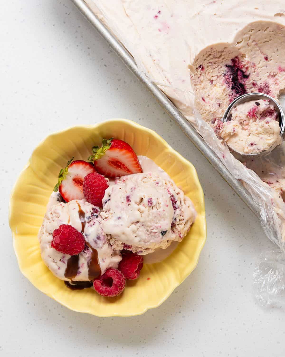 A yellow bowl with scoops of berry ice cream topped with fresh strawberries, raspberries, and chocolate sauce, next to a tray of ice cream with a scoop.