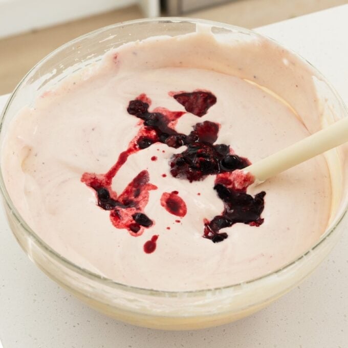 Berry Semifreddo A glass bowl filled with light pink batter being mixed with a spatula, with a swirl of dark berry sauce on top.