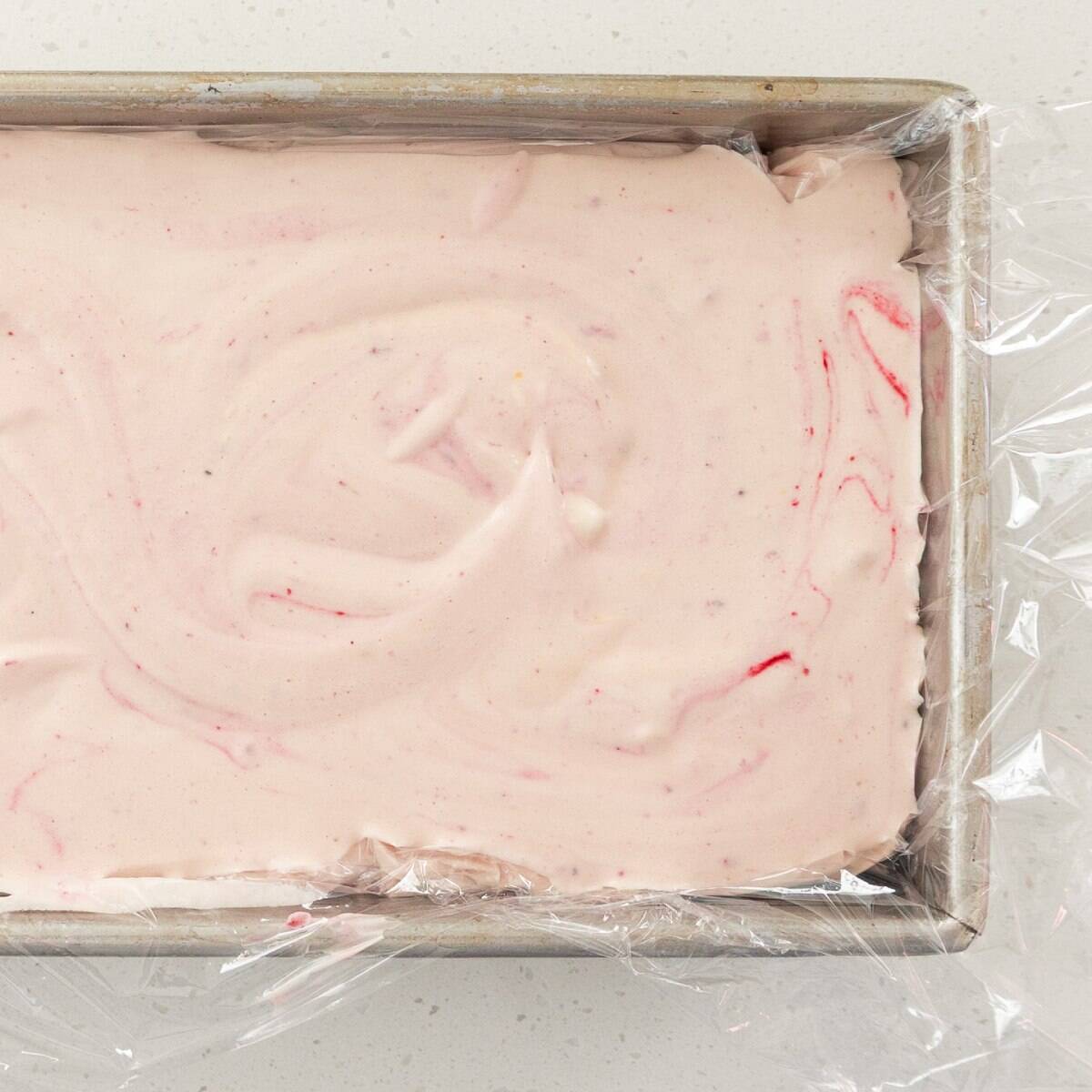 Berry Semifreddo A rectangular metal pan filled with pale pink, swirled ice cream mixture, partially covered with clear plastic wrap.