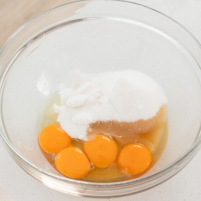 Berry Semifreddo A glass bowl containing four raw eggs, granulated sugar, and a small amount of white powder, likely baking powder or salt, on a white surface.