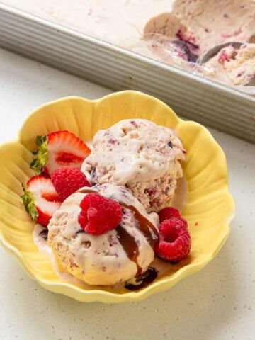 Two scoops of berry ice cream in a yellow bowl topped with chocolate sauce, garnished with raspberries and sliced strawberries. Ice cream tray with scoop in the background.