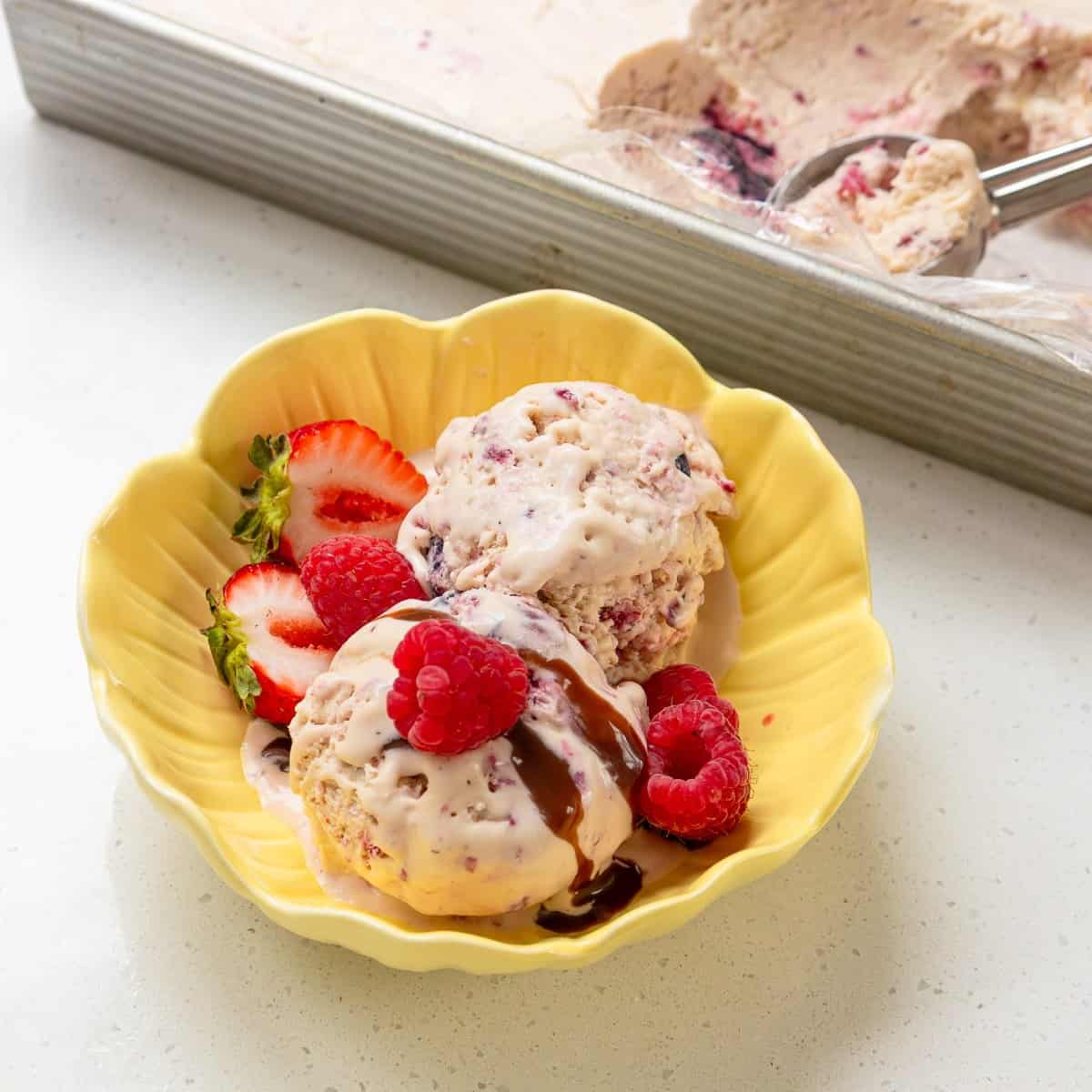 Berry Semifreddo Two scoops of berry ice cream in a yellow bowl topped with chocolate sauce, garnished with raspberries and sliced strawberries. Ice cream tray with scoop in the background.