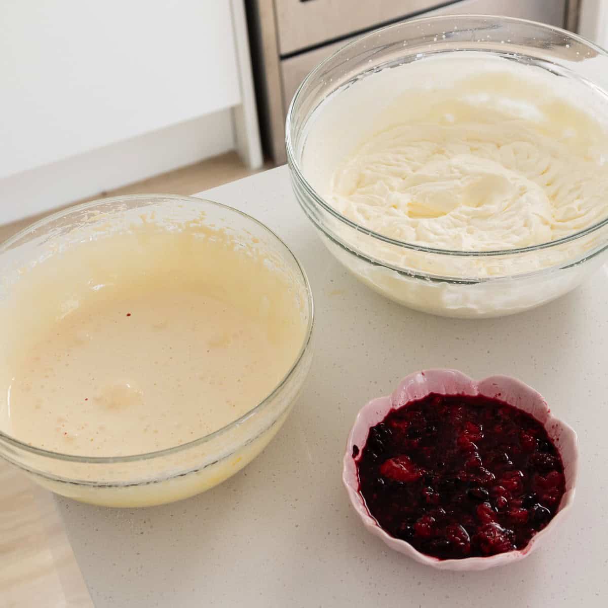 Berry Semifreddo Three bowls on a white countertop containing whipped cream, cake batter, and berry compote, prepared for baking or dessert assembly.