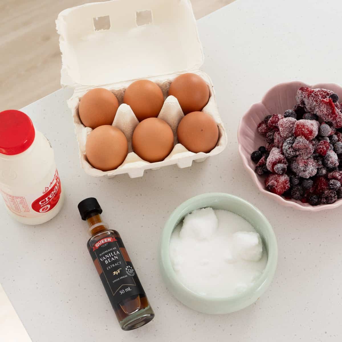 Eggs in a carton, a bottle of vanilla extract, cream, a bowl of sugar, and a bowl of frozen mixed berries on a white countertop.
