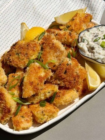 A plate of golden-brown breaded fish pieces garnished with green onions, served with lemon wedges and a bowl of creamy dipping sauce.