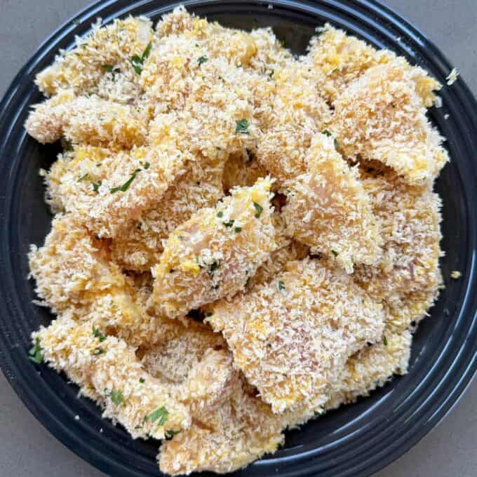 Crispy Panko Fish Bites A black plate filled with breaded, uncooked chicken nuggets coated in panko crumbs and sprinkled with herbs.