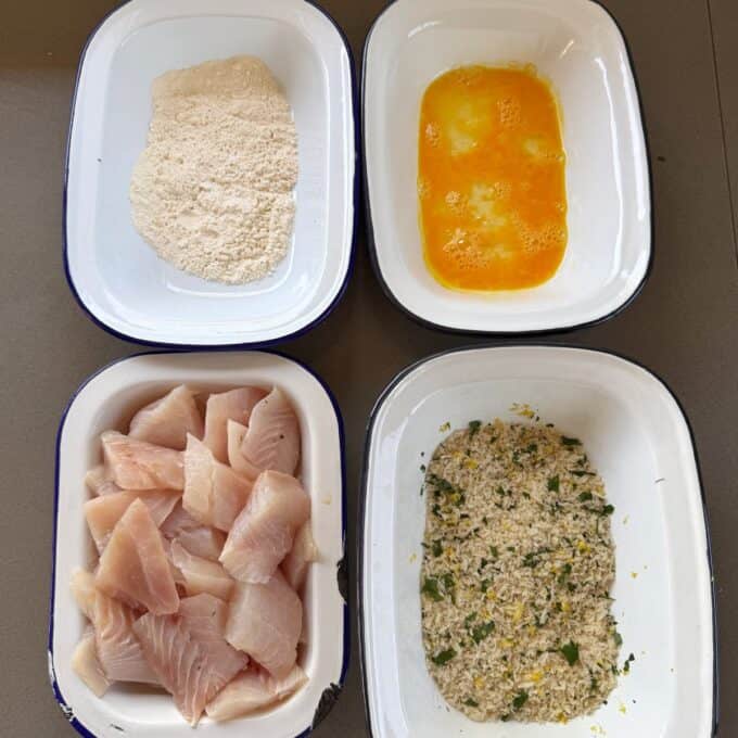 Crispy Panko Fish Bites Four dishes with ingredients for breaded fish: flour, beaten eggs, raw fish pieces, and a breadcrumb mixture with herbs and lemon zest.