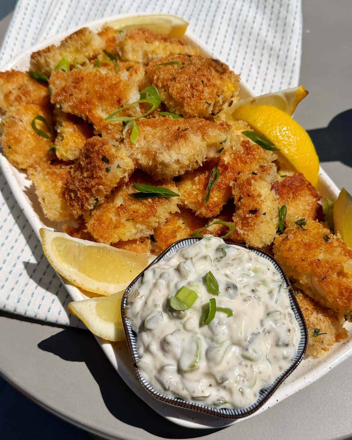 Crispy Panko Fish Bites A plate of breaded fried fish fillets garnished with green onions and lemon wedges, served with a bowl of creamy tartar sauce.