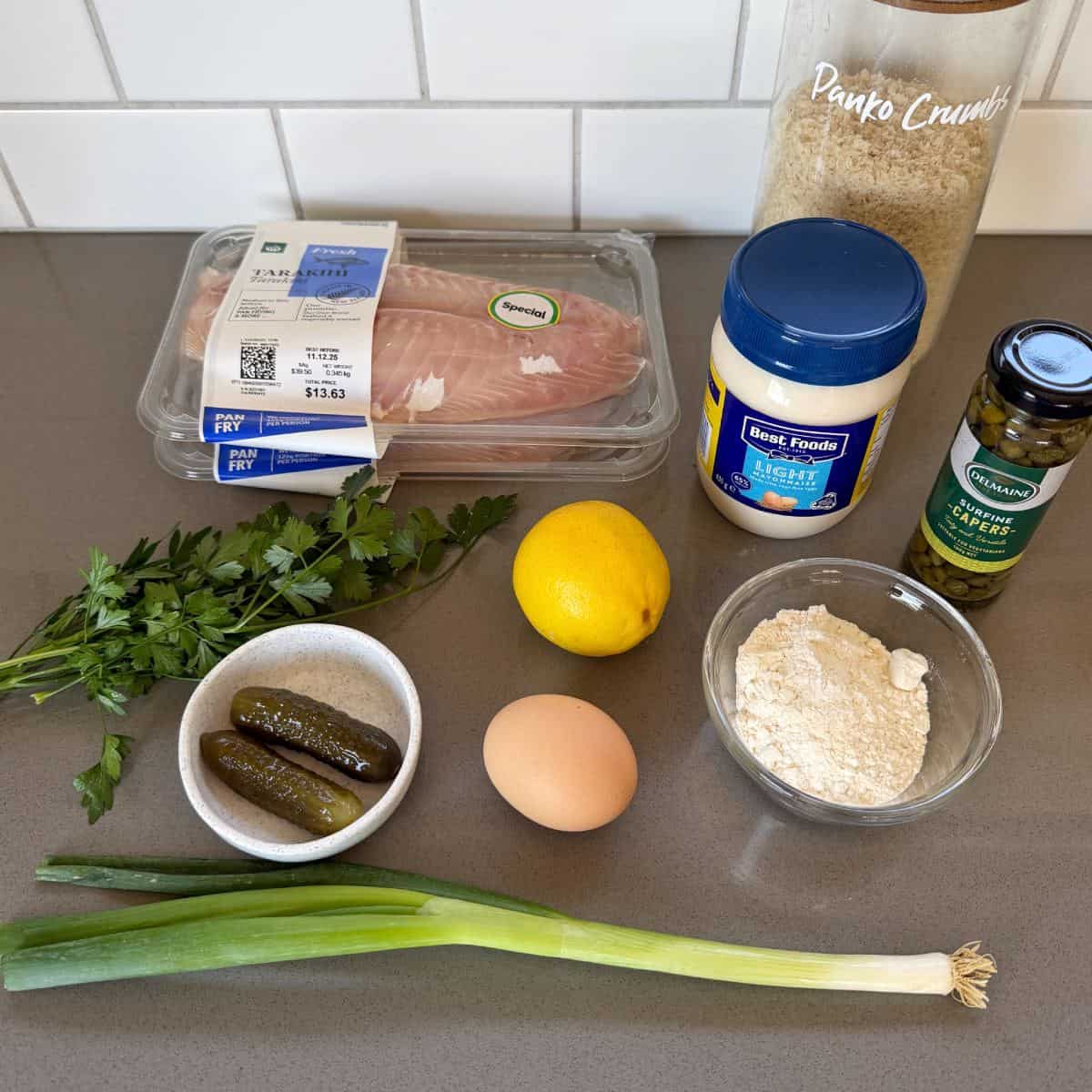Crispy Panko Fish Bites Ingredients on a countertop include raw fish fillets, panko crumbs, jarred mayonnaise, flour, green olives, pickles, parsley, a lemon, an egg, and green onions.