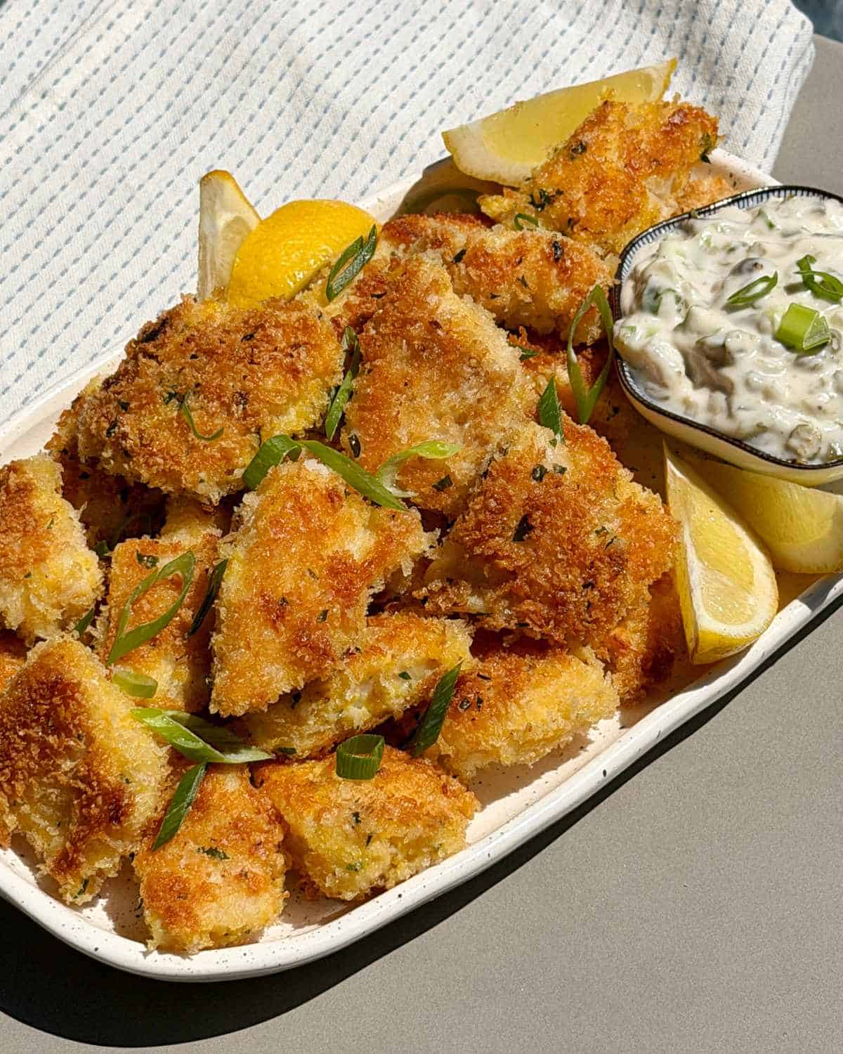 Crispy Panko Fish Bites A plate of breaded, baked fish pieces garnished with green onions and lemon wedges, served with a bowl of tartar sauce.