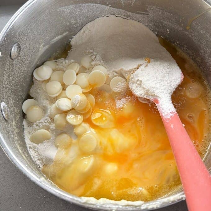 Feijoa and White Blondie A mixing bowl with flour, eggs, white chocolate chips, and a pink spatula, showing the beginning stages of baking preparation.