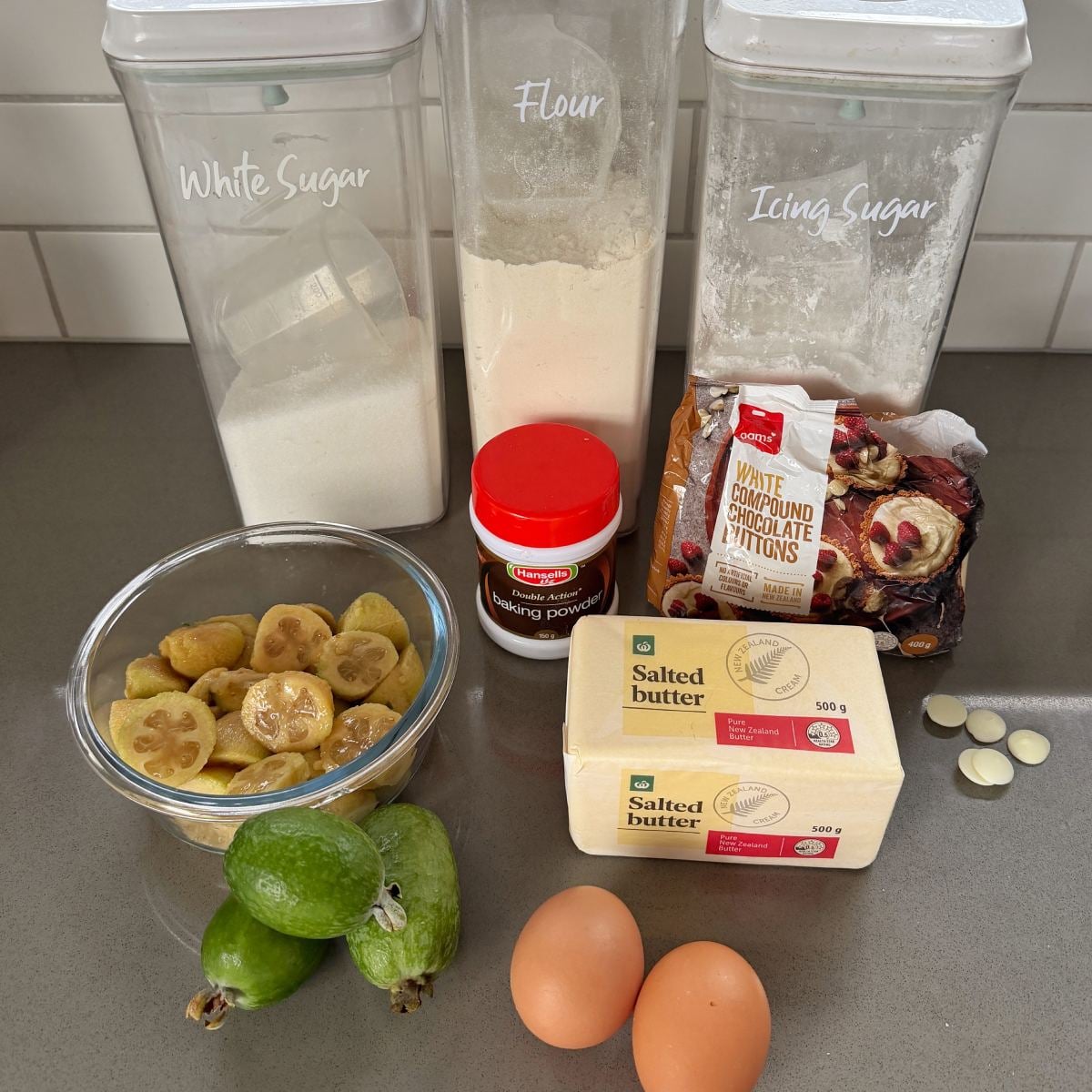 Feijoa and White Blondie Baking ingredients on a kitchen counter, including sugar, flour, icing sugar, baking powder, butter, white chocolate, dried figs, feijoas, eggs, and hazelnuts.